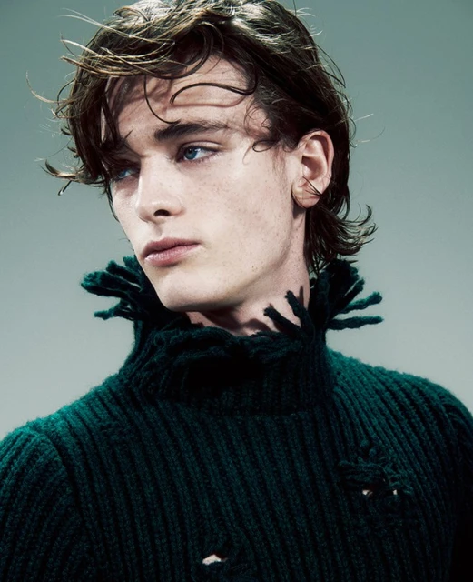 Close-up portrait of a young adult male with brown wavy hair and blue eyes wearing a green ribbed knit turtleneck sweater indoors.