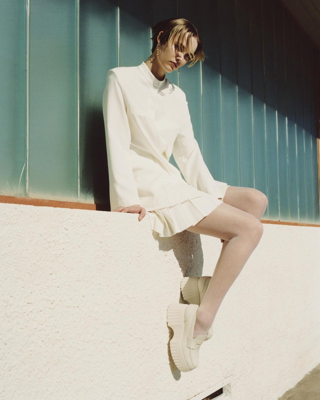 A woman sits on a ledge outdoors wearing a cream-colored dress and platform shoes in a fashion editorial shot against a window wall.