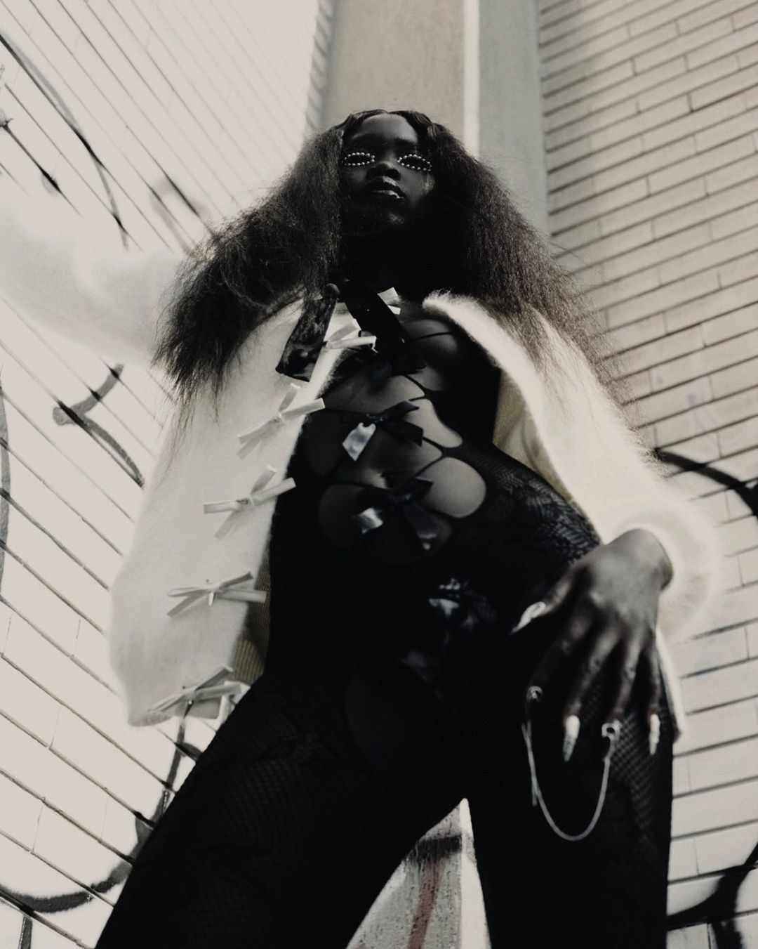 A striking portrait of a black female adult fashion model posing in front of a graffiti wall. She is wearing a white sweater with a black lace bodysuit.