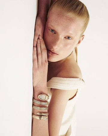 Close-up portrait of a woman wearing a coiled gold serpent watch on her wrist against a white background