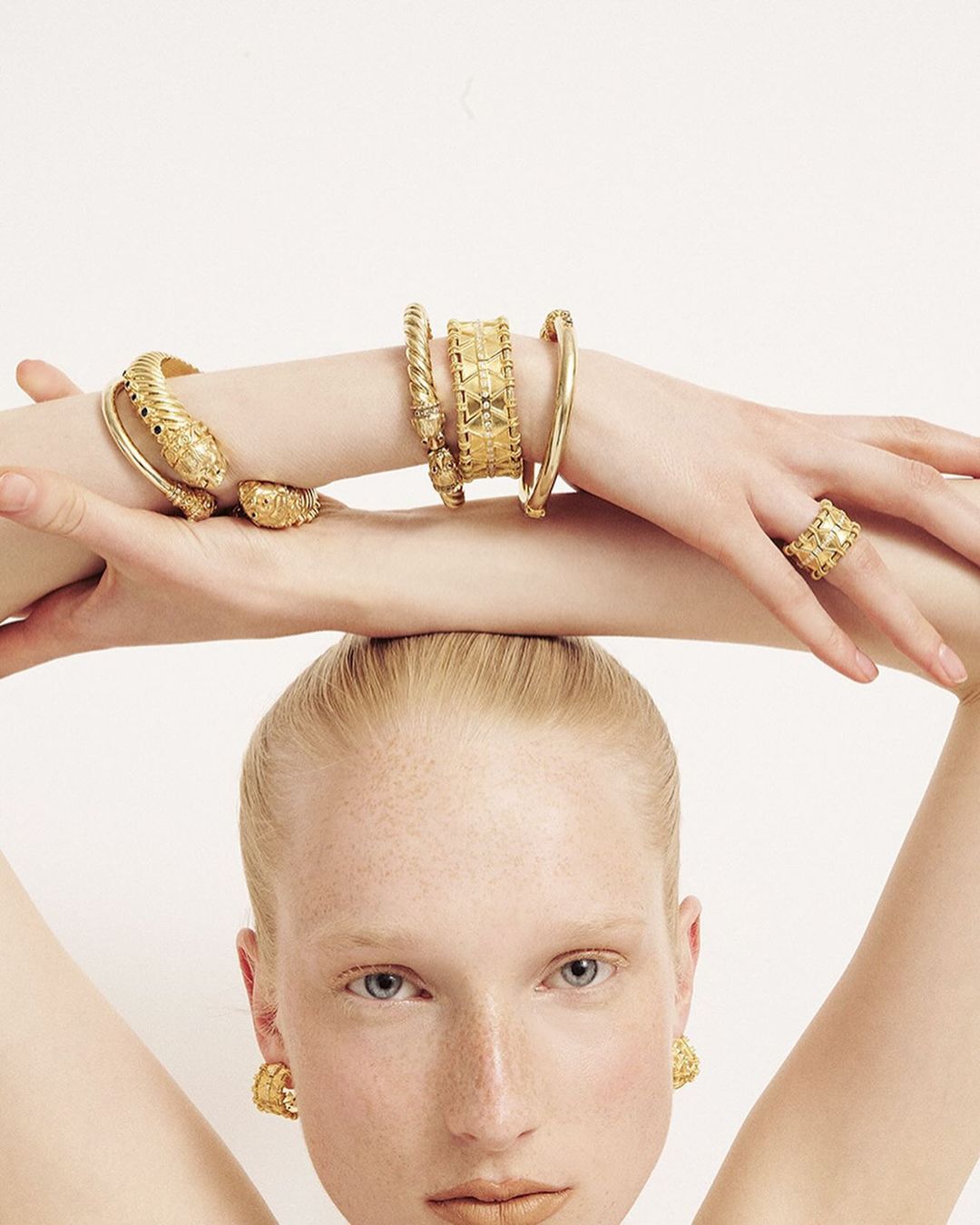 Close-up shot of a fair-skinned adult woman wearing golden jewelry, including a snake bracelet, rings, and statement earrings, on a white background.