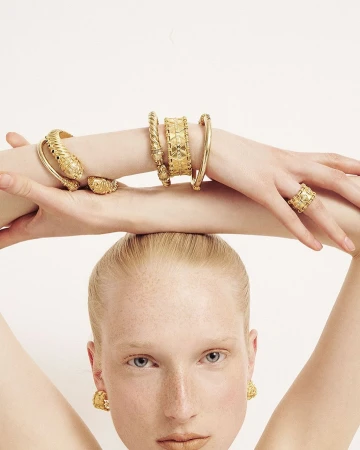 Close-up portrait of a model with arms raised showcasing ornate gold bracelets and matching rings against a plain white background.