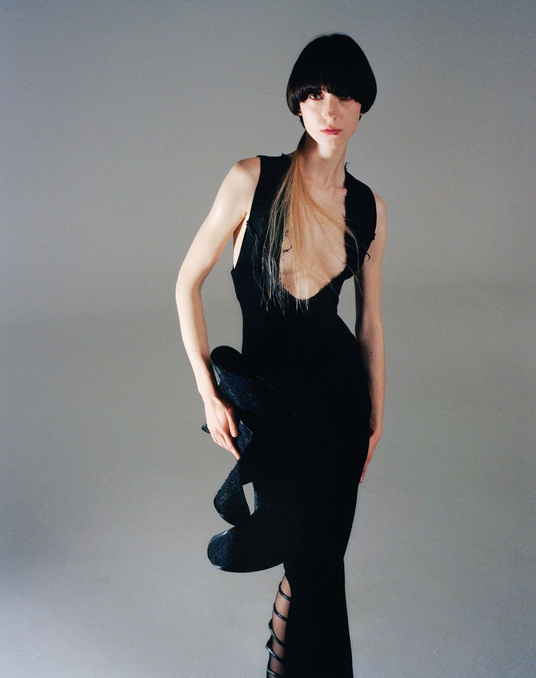 A slender fashion model poses in a sleeveless black gown with cut-out details against a neutral gray background, highlighting the dress's design.