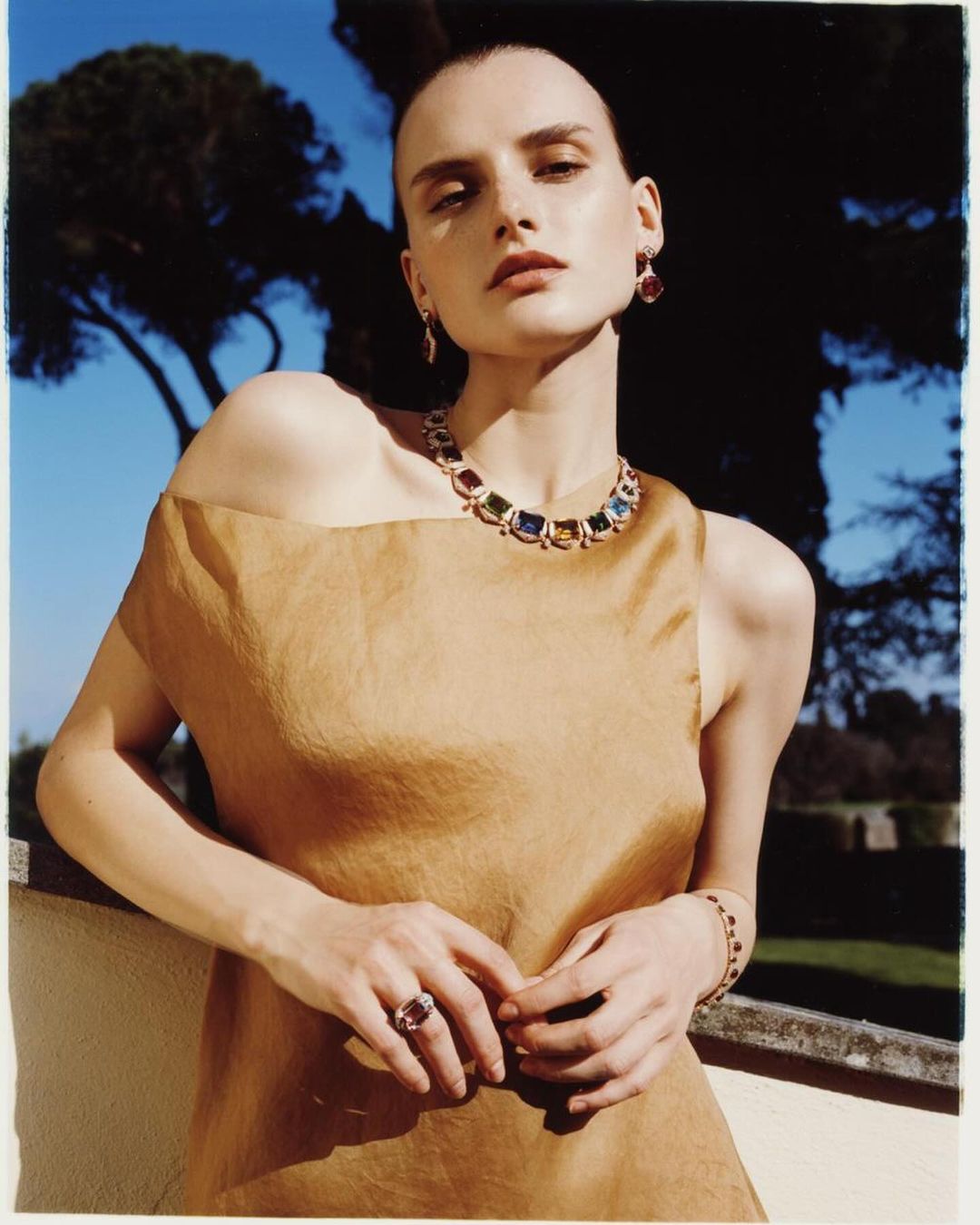 Elegant woman in tan dress poses outdoors, adorned with sapphire jewelry set including necklace, earrings, ring, and bracelet.