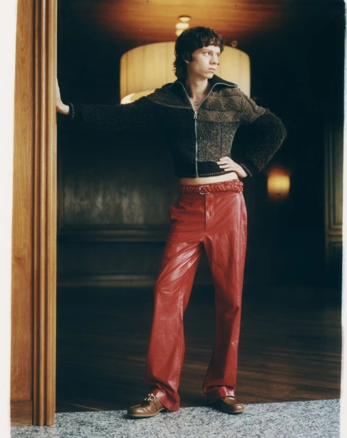 An adult male poses in red leather pants and a knit top in a doorway, looking to his right, with warm lighting in the background.