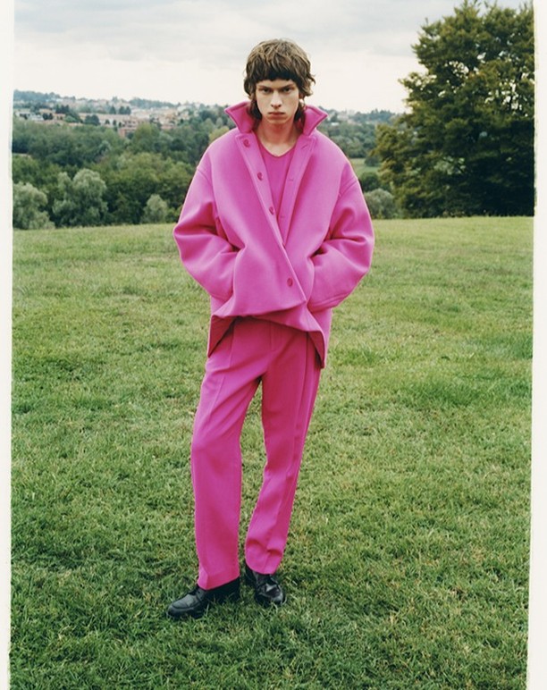 Man poses outdoors in vibrant pink coat and matching pants on green grass, showcasing a fashion-forward editorial style with natural light.