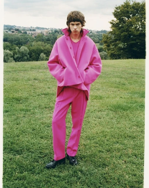 Man poses outdoors in vibrant pink coat and matching pants on green grass, showcasing a fashion-forward editorial style with natural light.