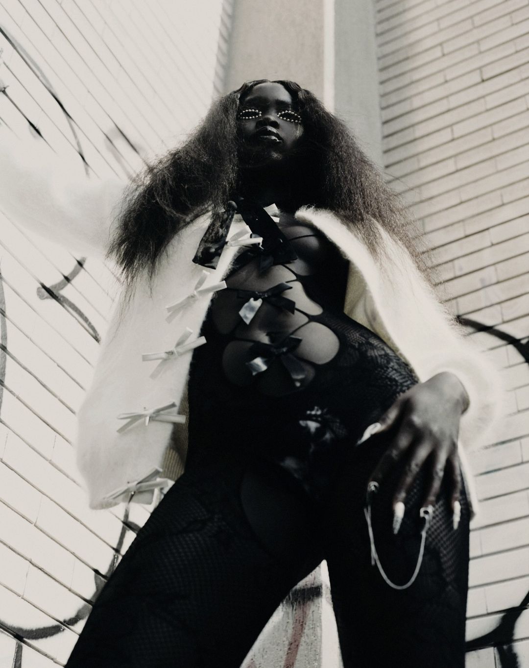 A dark-skinned woman with long dark hair poses in a black lace outfit and a white cardigan with bows, against a graffiti wall.