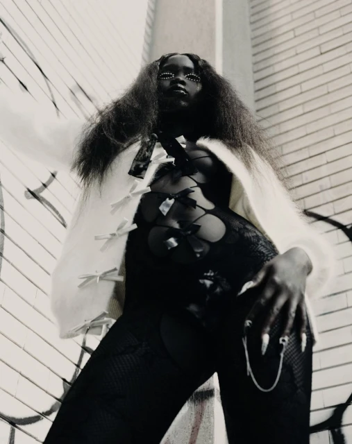 A dark-skinned woman with long dark hair poses in a black lace outfit and a white cardigan with bows, against a graffiti wall.