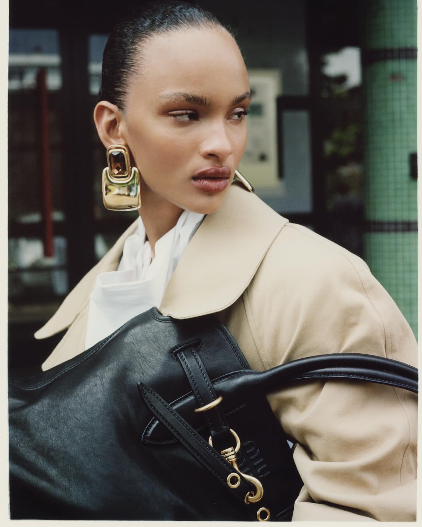 Model wearing large gold earrings and a beige coat holding a black leather bag, photographed by Antonio Dicorato in Paris