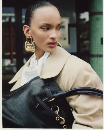 Model wearing large gold earrings and a beige coat holding a black leather bag, photographed by Antonio Dicorato in Paris