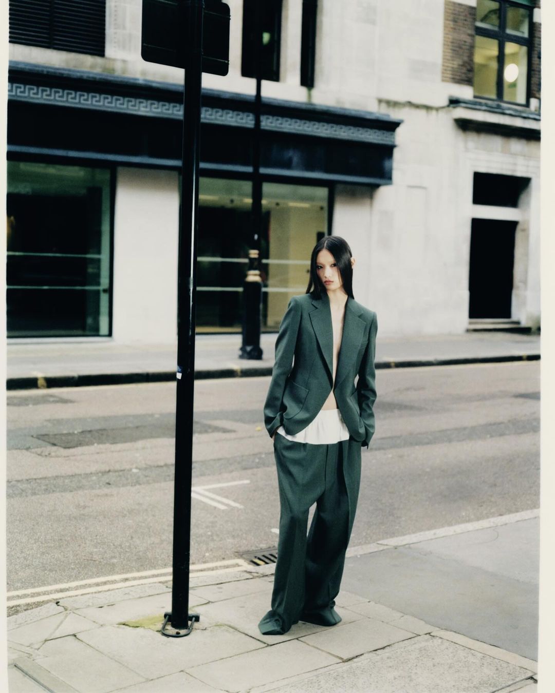 Fashion shot of a young adult woman posing in an oversized grey-green suit on a city sidewalk with buildings in the background.