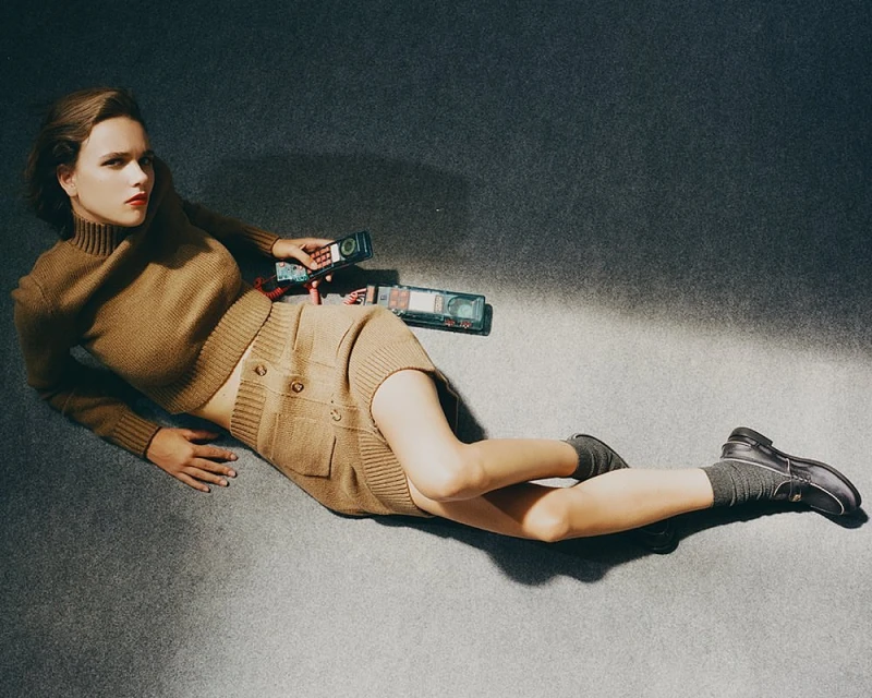 Woman in tan sweater and skirt poses on gray floor with vintage cell phones, facing the camera in a fashion editorial shot.