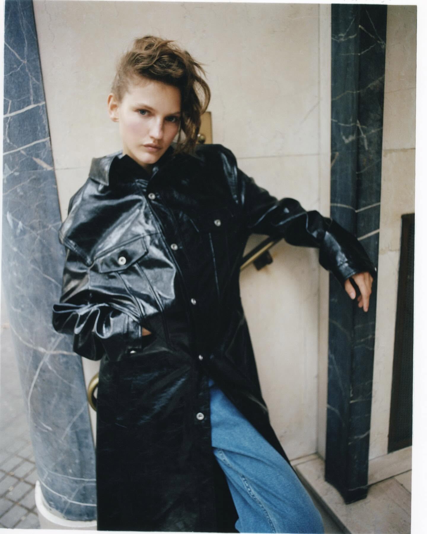 Fashionable young adult posing in a black vinyl trench coat and jeans leaning against a marbled column in an urban setting.