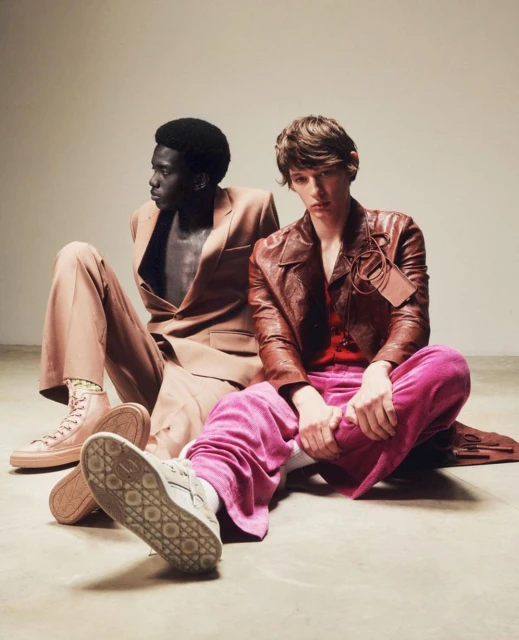Two stylish male models pose seated in a studio wearing suits, one in light brown and the other in a brown leather jacket and pink pants.