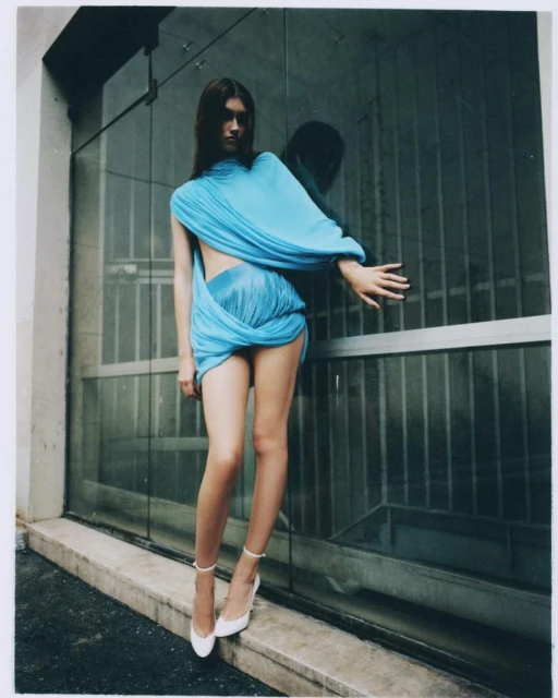 Young woman in a turquoise draped dress and white heels poses in an urban setting with a textured window behind her.