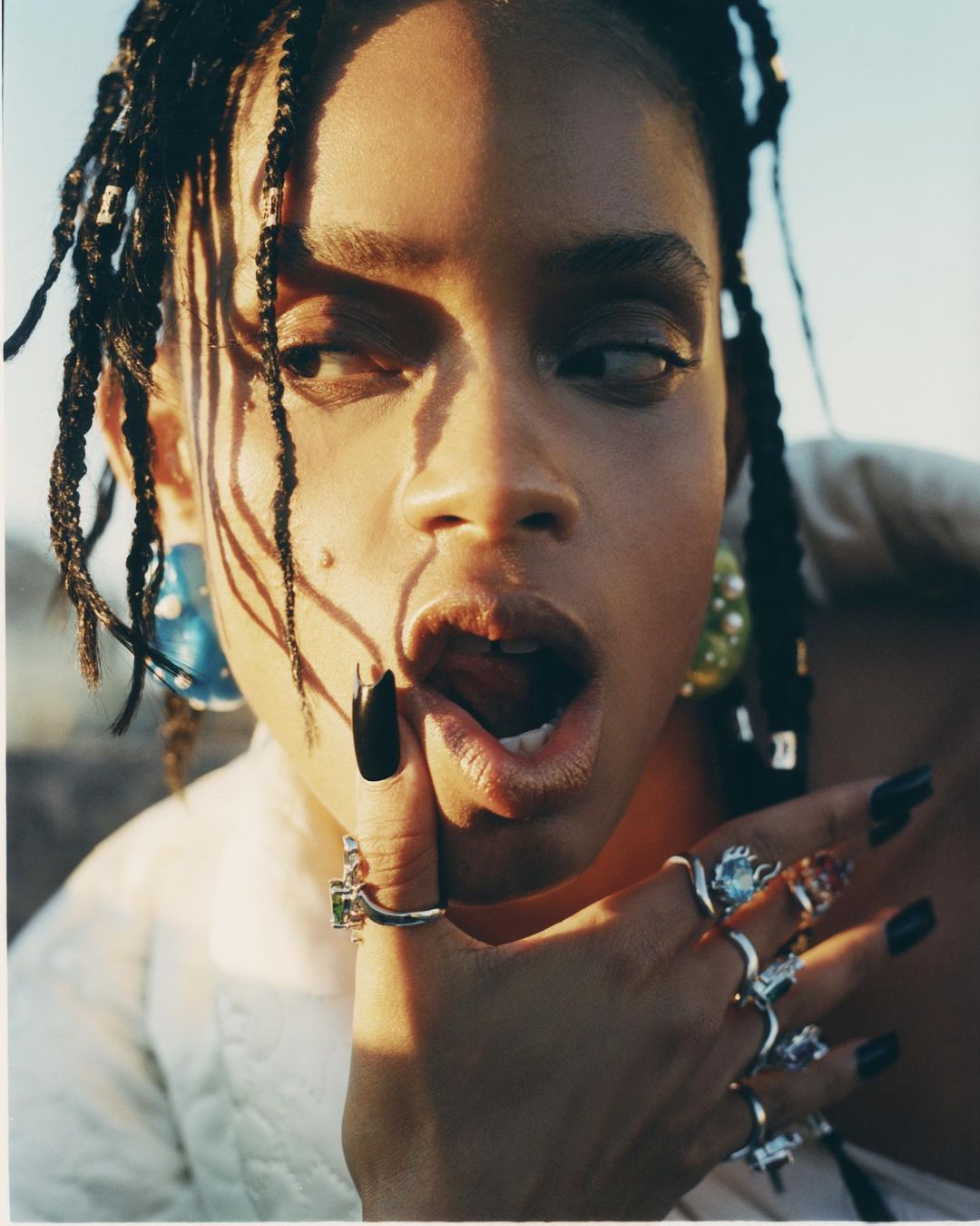 Close-up portrait of a woman with braided hair and dark manicure, wearing rings, with her tongue slightly out, against a bright background.