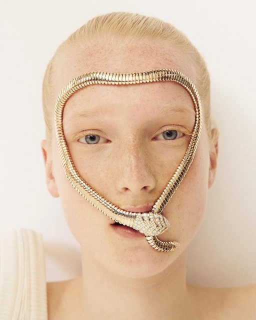 Woman with fair skin and blonde hair, wearing a gold snake-shaped necklace with diamond head, against a neutral white background.