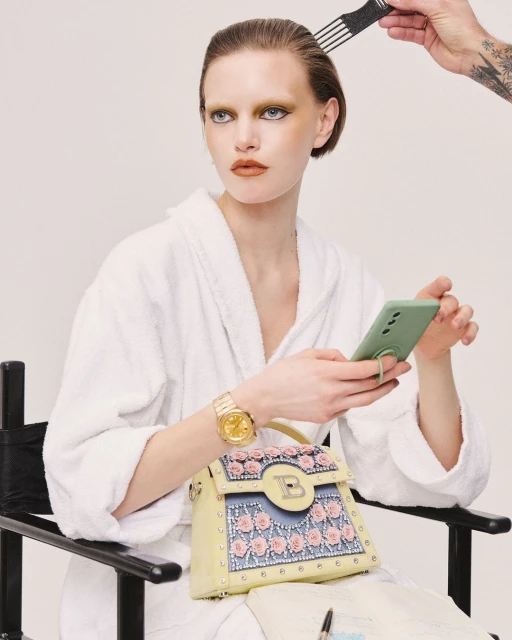 A model in a white robe is sitting on a black chair, using a phone, with a designer purse next to her and a stylist fixing her hair.