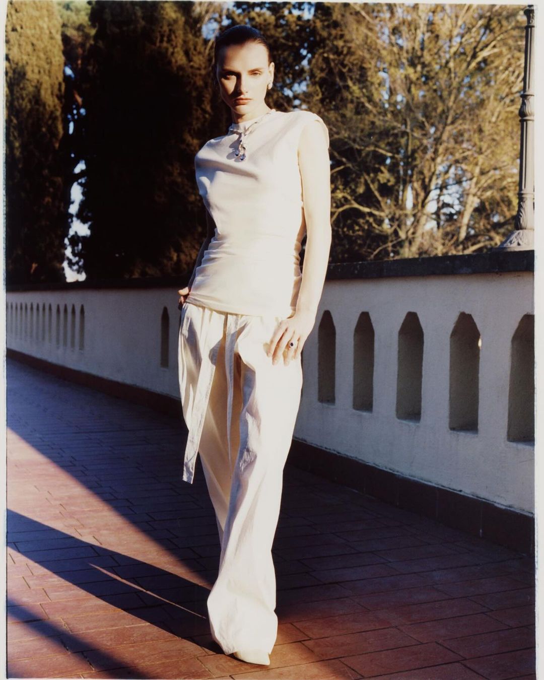 Fashion shot of a slim woman with dark hair wearing a cream top and pants, standing on a brick surface with a tiled wall and trees in the background.