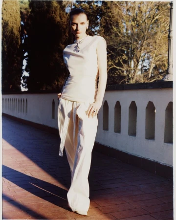 Fashion shot of a slim woman with dark hair wearing a cream top and pants, standing on a brick surface with a tiled wall and trees in the background.