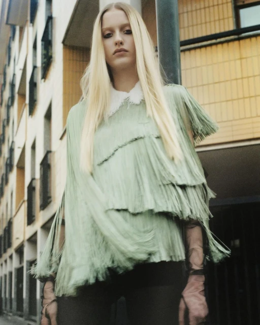 Fashion shot of a blonde adult woman wearing a sage green fringed top with a white collar against an urban exterior background.