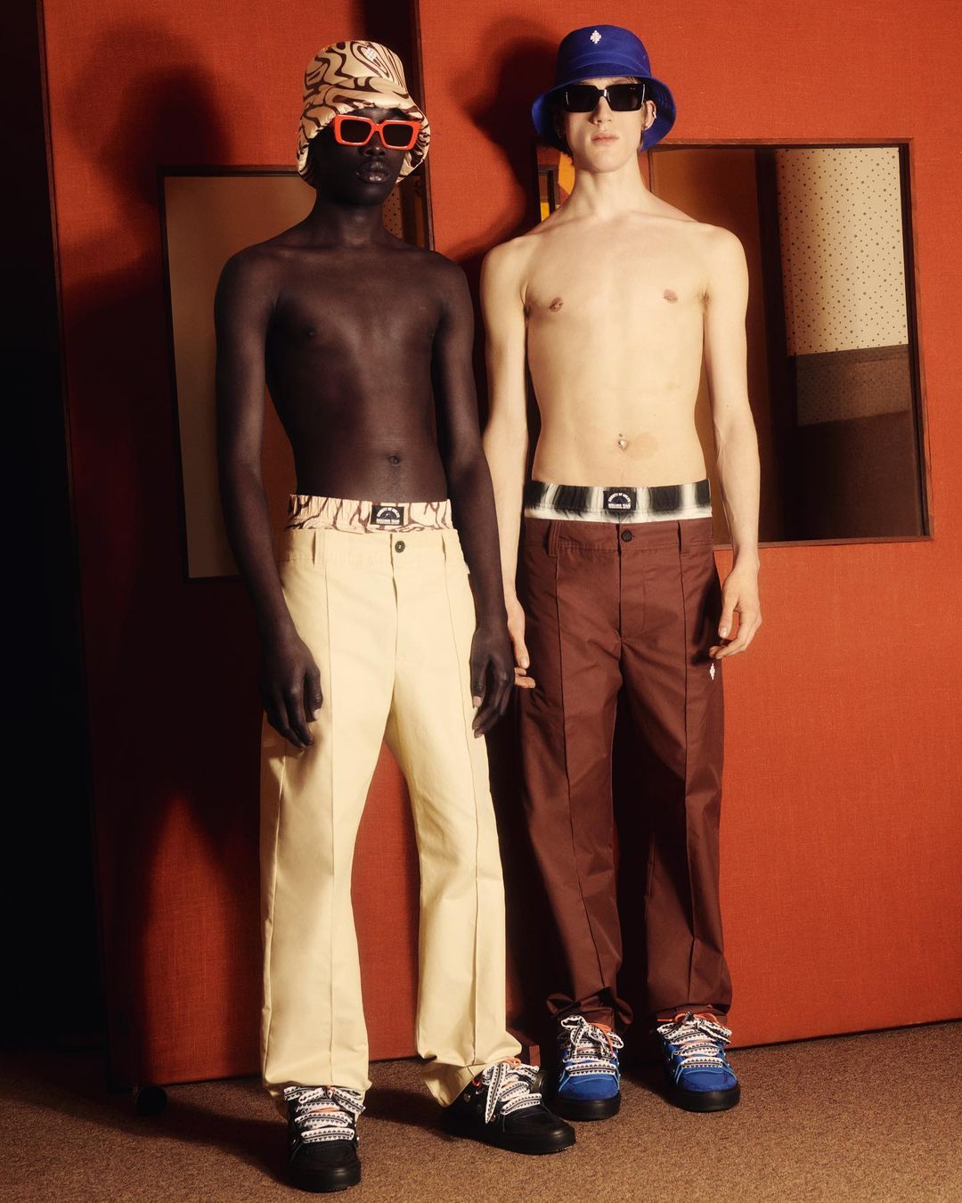 Two shirtless male fashion models in bucket hats, belted trousers, and sneakers pose indoors against a terracotta-colored wall and doorway.