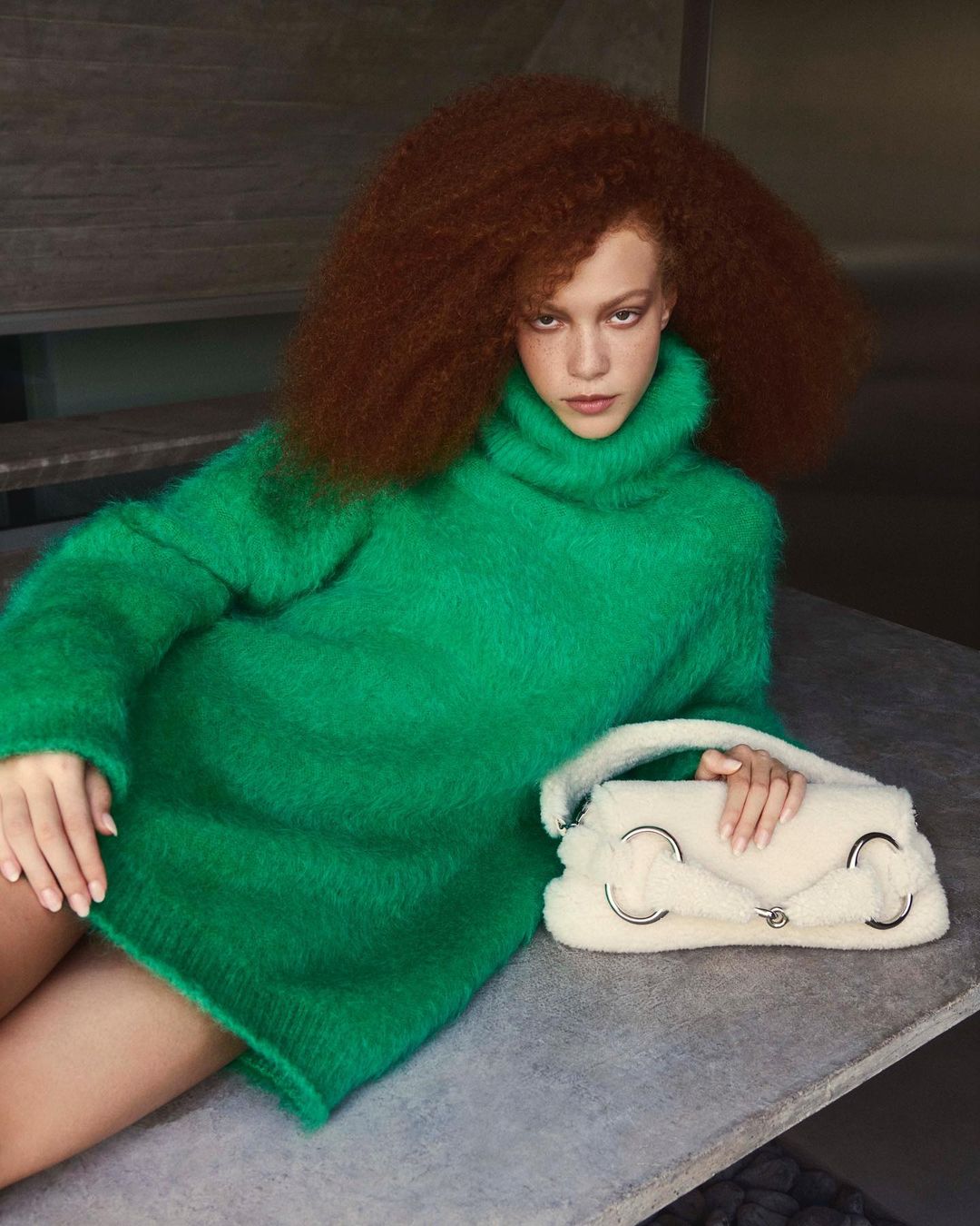 A young red-haired woman in a green sweater dress poses on a concrete structure with a white furry bag in a fashion setting.