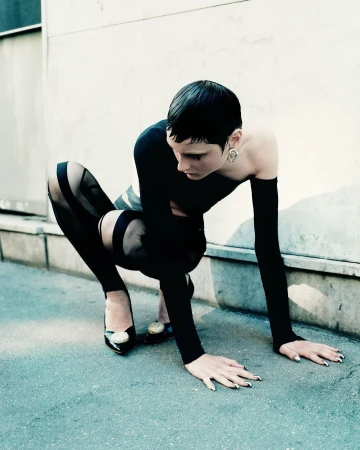 Model with short dark hair crouched on pavement wearing a black top and heels with metallic toe embellishments