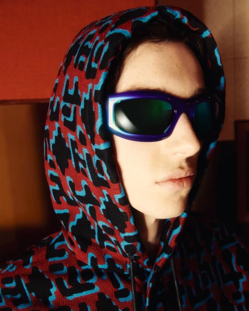 Profile shot of a person with fair skin wearing geometric sunglasses and a hooded sweater, with an indoor setting in the background.