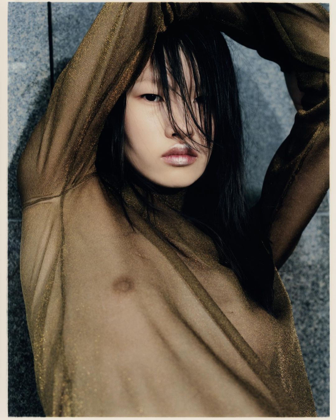 An Asian woman poses with arms raised, wearing a sheer gold top against a textured grey wall, emphasizing fashion and portraiture.