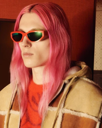A fair-skinned adult male with pink hair and orange sunglasses poses indoors, wearing a patterned jacket and an orange sweater.