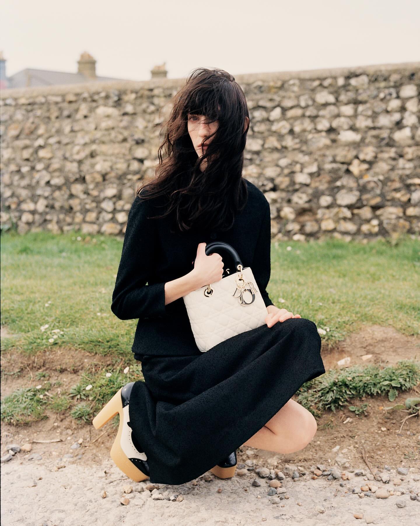 Adult female model in black dress kneeling outside, holding cream-colored quilted handbag with black handle and platform heels by stone wall.