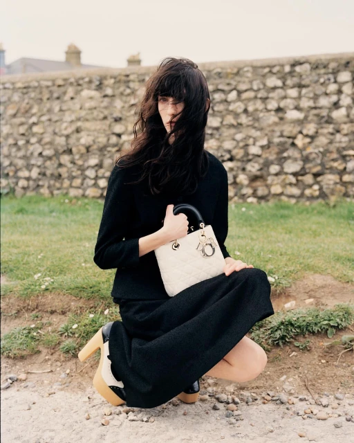 Adult female model in black dress kneeling outside, holding cream-colored quilted handbag with black handle and platform heels by stone wall.