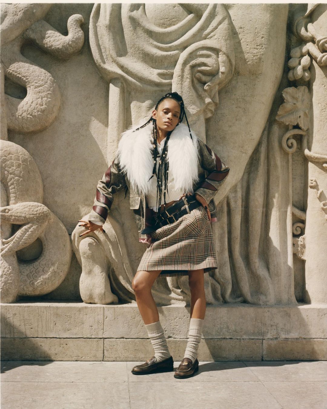 A young adult woman with braided hair wearing a skirt and jacket poses outdoors against a detailed stone wall with light shadows.