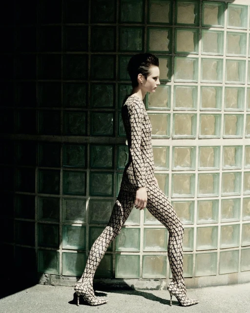 A slender adult female walking in a printed full bodysuit and high heels, captured in an outdoor shot against a textured glass brick wall.