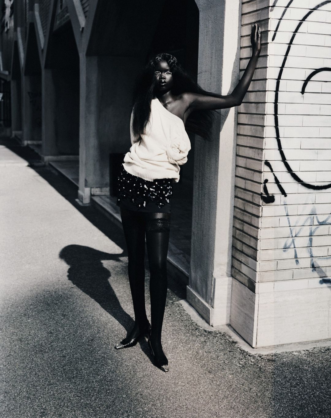 Elegant Black woman in fashion attire poses in an urban outdoor setting, wearing a cream sweater, black skirt, and black stockings.