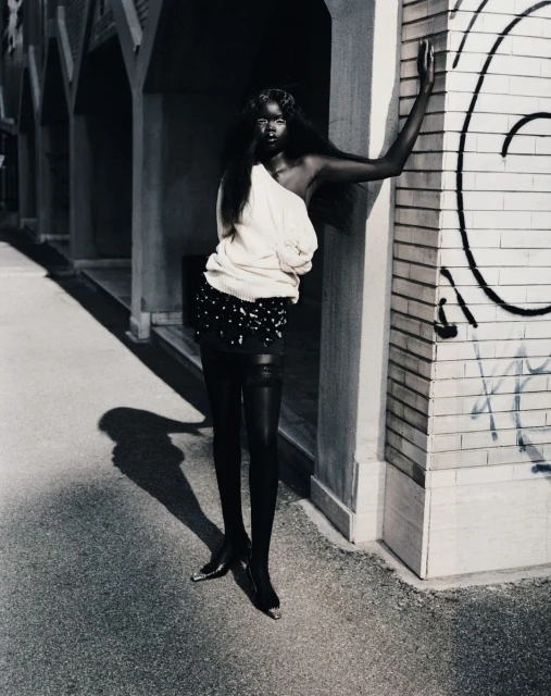 Elegant Black woman in fashion attire poses in an urban outdoor setting, wearing a cream sweater, black skirt, and black stockings.