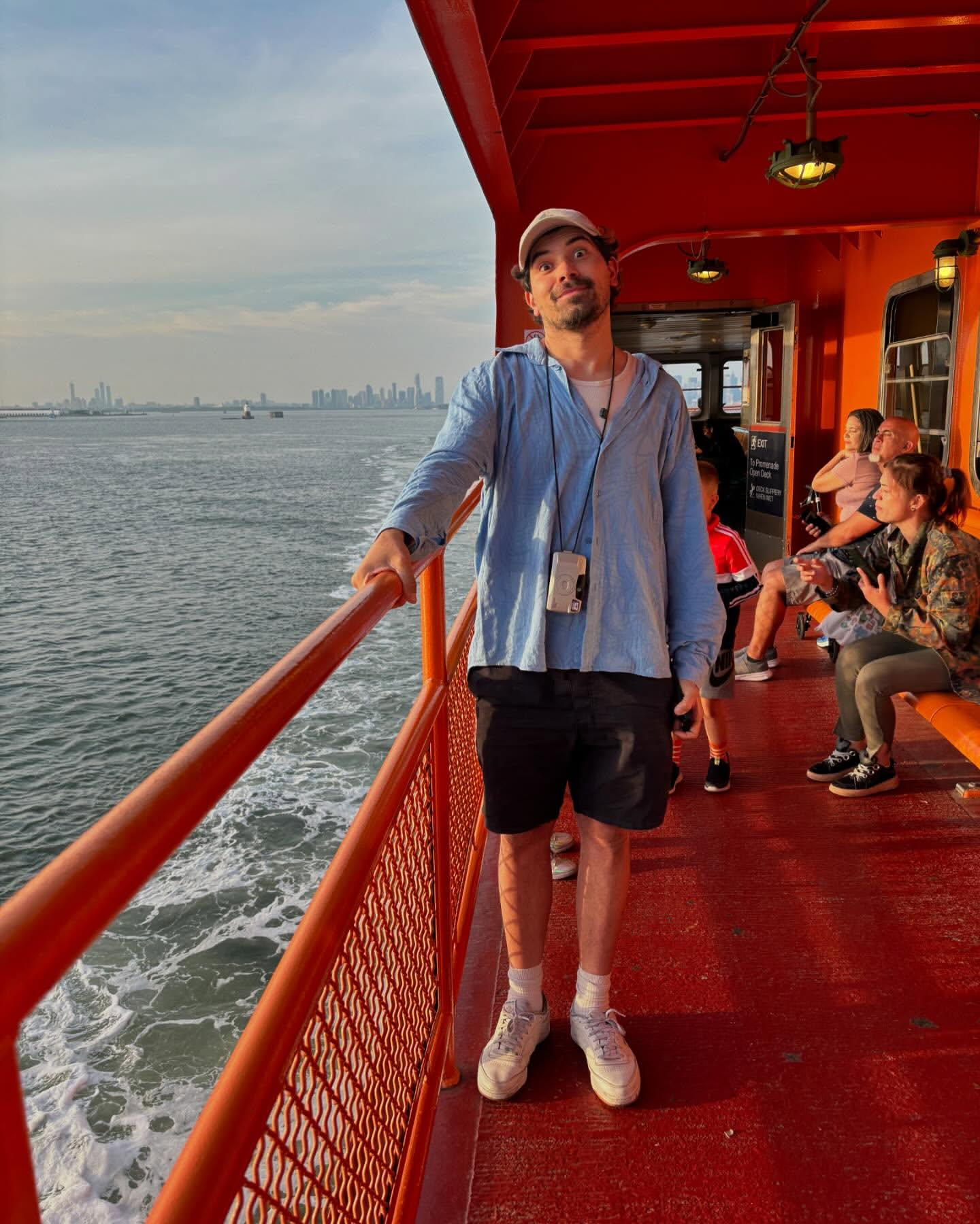 Man stands on orange ferry railing with ocean and cityscape in background. He wears a blue shirt, black shorts, and a baseball cap.