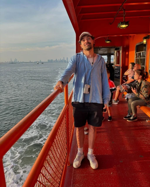 Man stands on orange ferry railing with ocean and cityscape in background. He wears a blue shirt, black shorts, and a baseball cap.