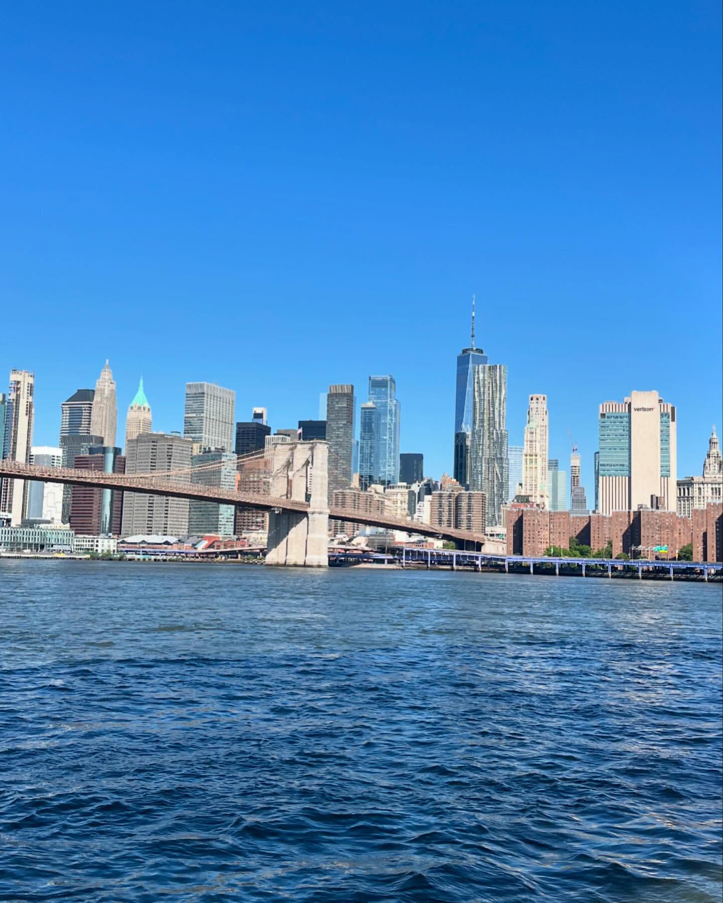 New York City skyline featuring the Brooklyn Bridge and skyscrapers, set against a bright blue sky with water in the foreground.