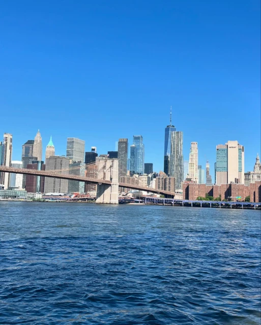 New York City skyline featuring the Brooklyn Bridge and skyscrapers, set against a bright blue sky with water in the foreground.