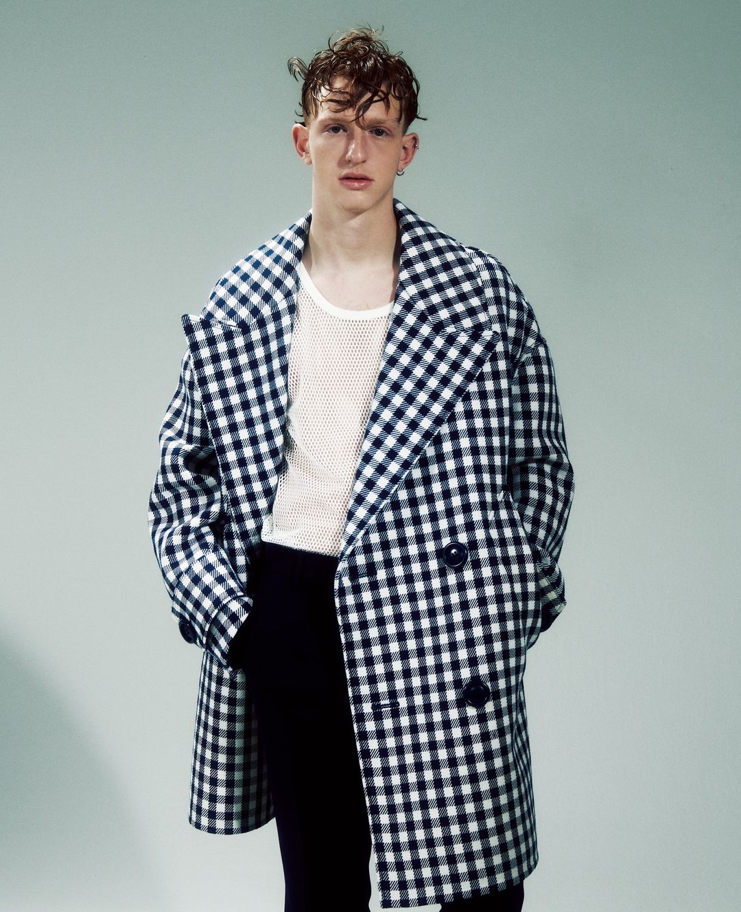 A young adult male poses in a studio wearing a gingham overcoat, textured white top, and dark pants against a light background.