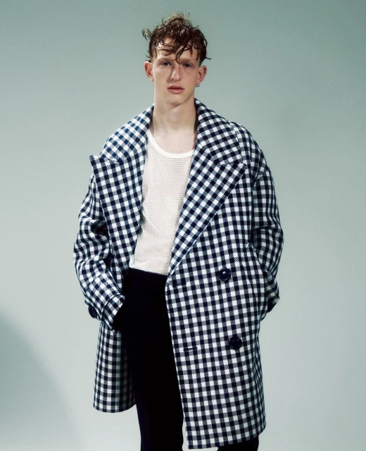 A young adult male poses in a studio wearing a gingham overcoat, textured white top, and dark pants against a light background.