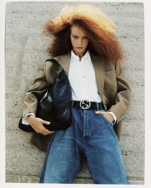 A woman with voluminous red curly hair poses in a studio, wearing a tan blazer, white shirt, jeans, and a Gucci belt, holding a black bag.