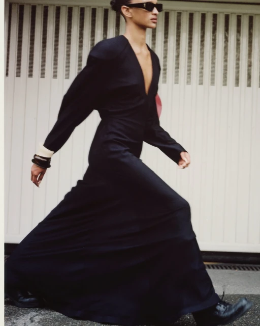 Woman in a stylish black dress and sunglasses walking on an urban street in a fashion pose, showcasing fashion editorial style.