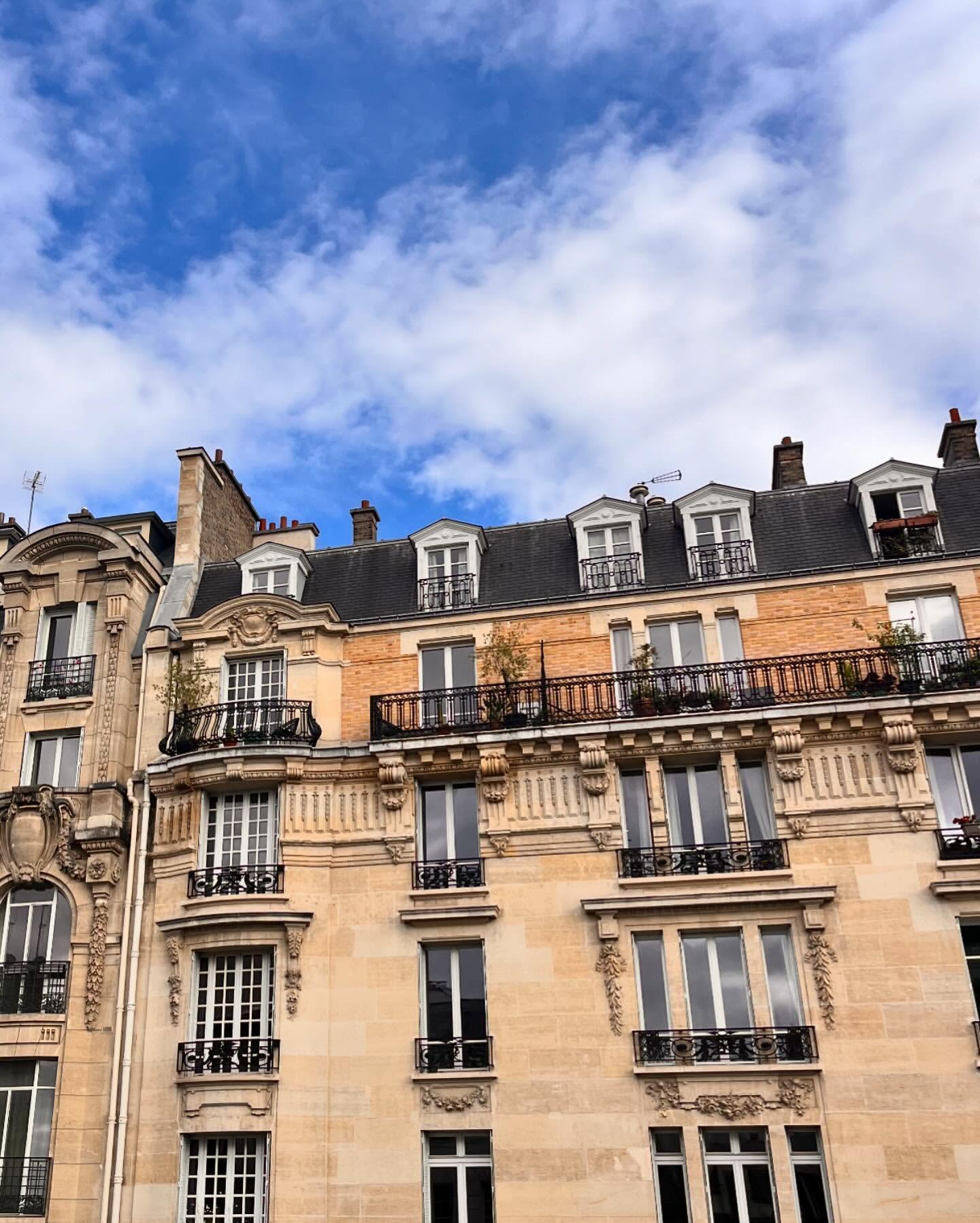 Exterior view of Parisian architecture showing sandstone apartment buildings with black metal balconies under a blue sky with clouds.