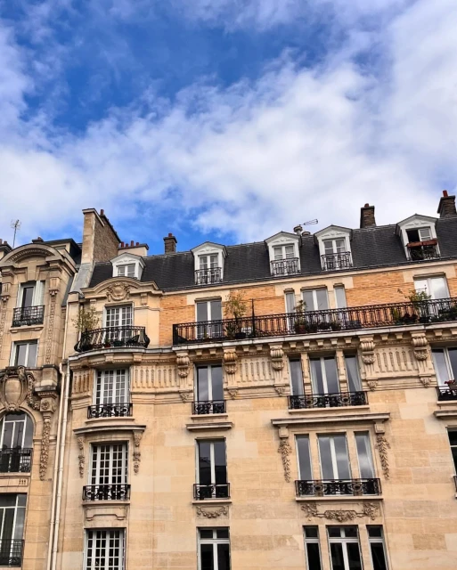 Exterior view of Parisian architecture showing sandstone apartment buildings with black metal balconies under a blue sky with clouds.