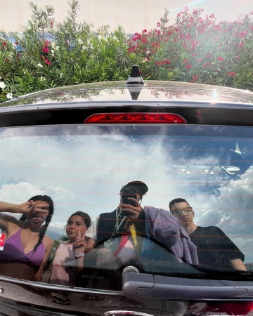 Group of four people reflected in the back of a black car, with red and white flowers and greenery in the background under a bright sky.