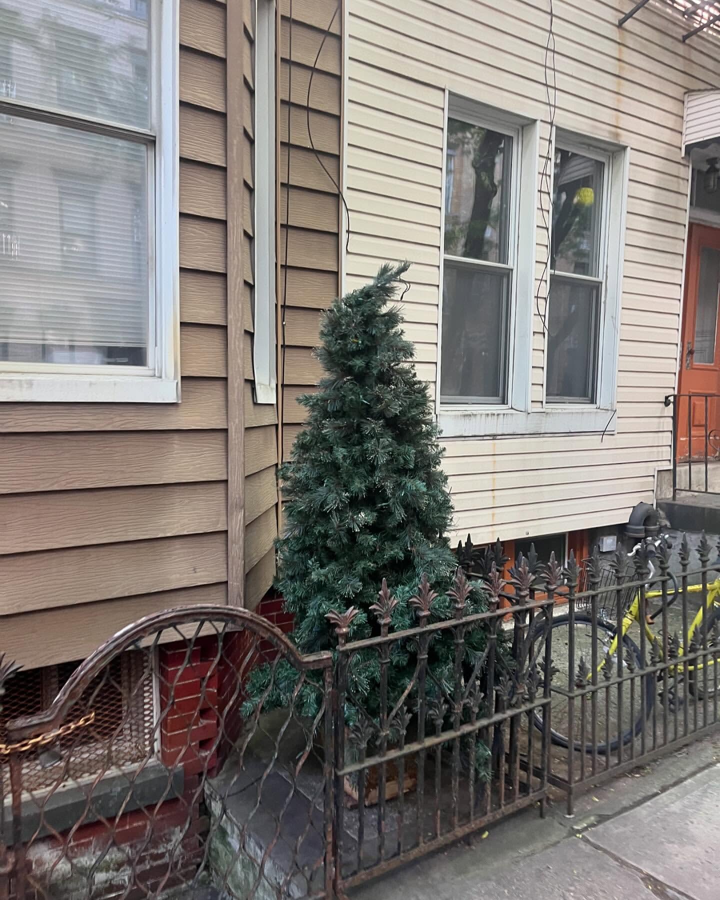 Artificial Christmas tree outside a building with beige siding and white trim, near a wrought iron fence and a parked yellow bicycle.